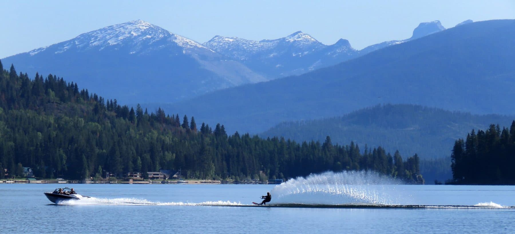 A water-skier carves across Priest Lake beneath forested hills and the snow-capped Selkirk Mountains.